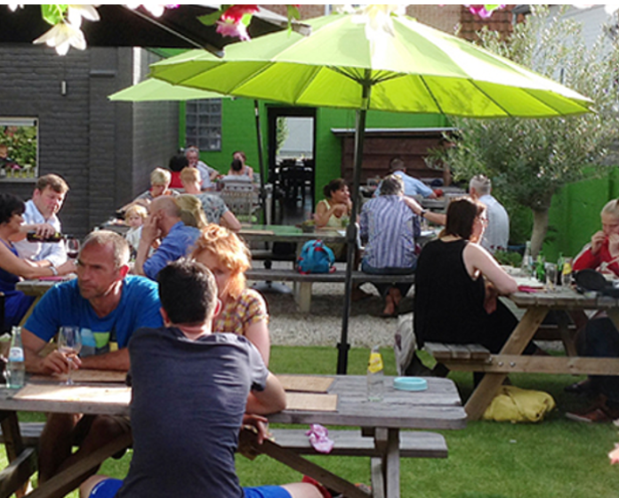 Gezellig buitenterras met picknicktafels, een groene parasol en gasten die samen zitten te eten in zonnig weer