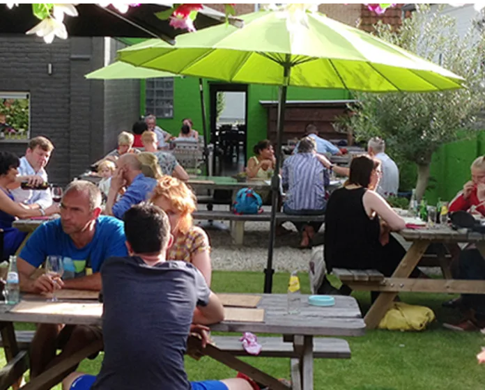 Gezellig buitenterras met picknicktafels en een groene parasol, gasten aan het tafelen in de zon